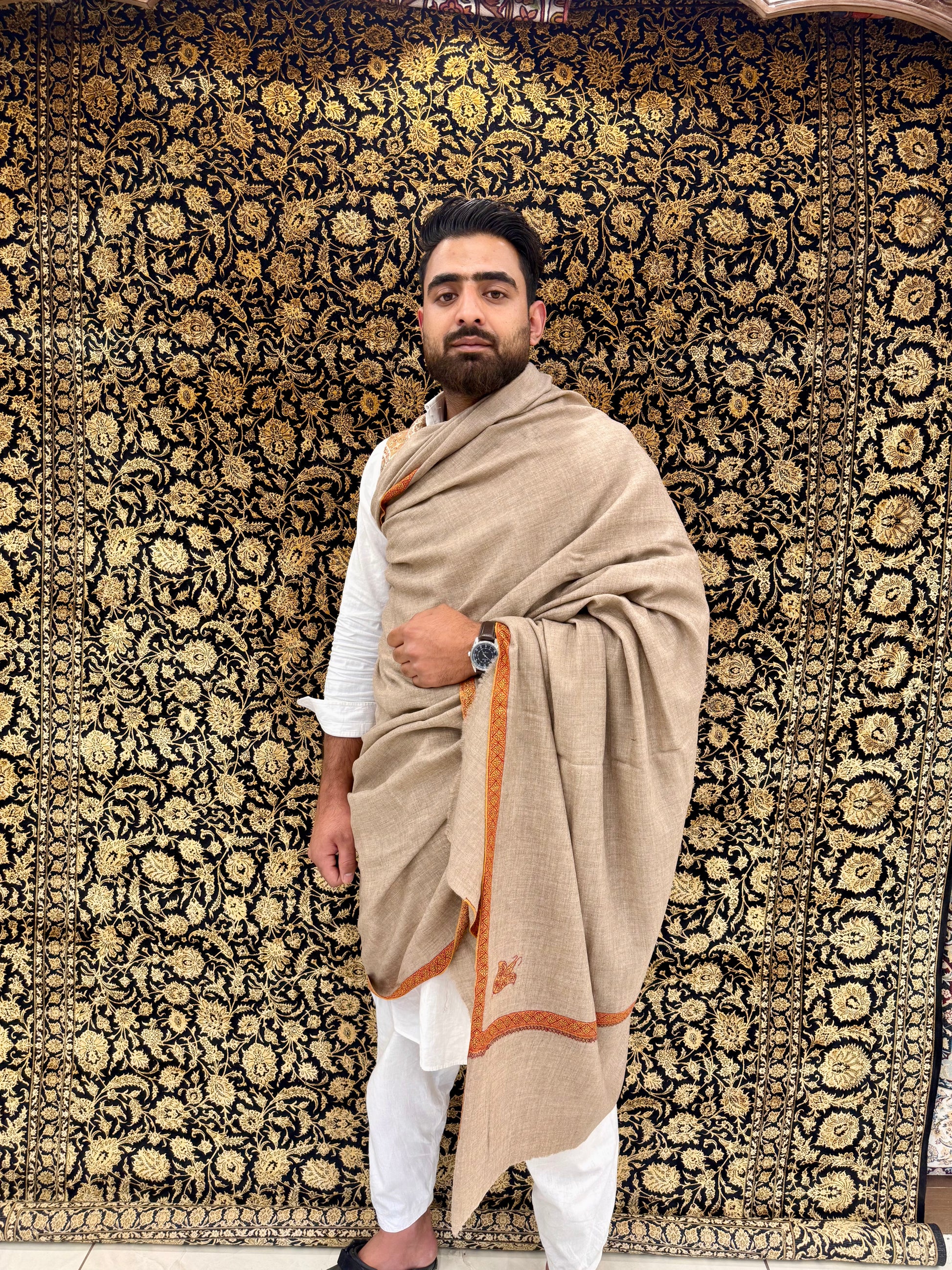 Man wearing a beige shawl with orange border against a patterned wall.